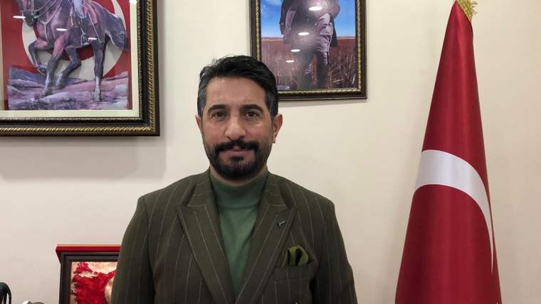 Kısa sakallı bir adam, yeşil bir tişört ve çizgili bir ceket giymiş, arkasında Türk bayrağı ve çerçevelenmiş fotoğraflar var.