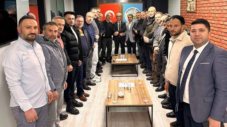 İç mekânda, ortadaki sehpaların çevresinde duran çok sayıda kişi birlikte poz veriyor.