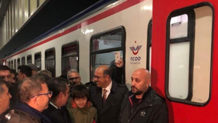 Kırmızı-beyaz tren vagonunun yanında kalabalık bir grup, aralarında elinde çiçek tutan bir çocukla görülüyor.