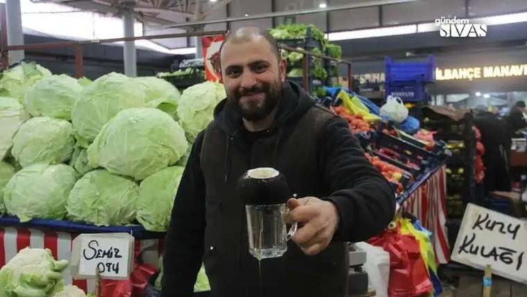 Kapalı pazarda sakallı bir kişi, elinde bardak tutarken yanında lahanalar ve sebze tezgâhları görülüyor.