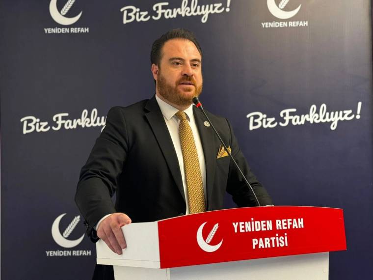 Mikrofon başında konuşan bir kişi, üzerinde Yeniden Refah Partisi yazan kürsüde duruyor.