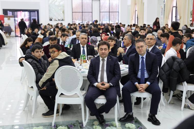 Bir etkinlikte oturan gençler ve yetişkinler, yemek masalarının etrafında toplanmış.