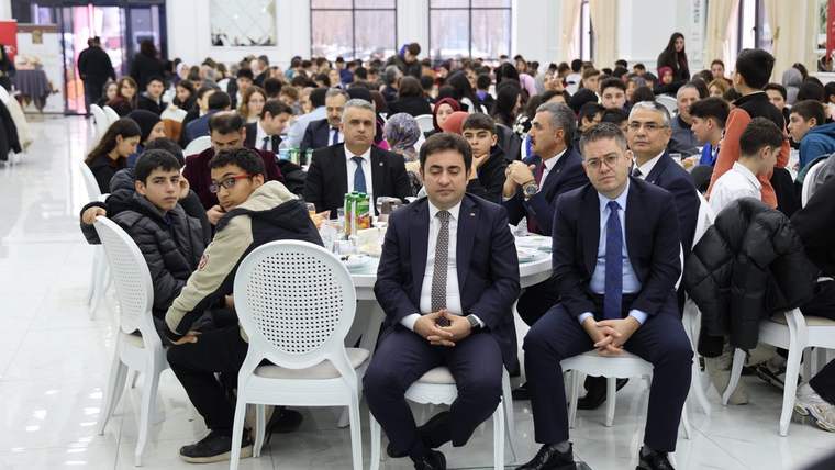 Bir etkinlikte oturan gençler ve yetişkinler, yemek masalarının etrafında toplanmış.