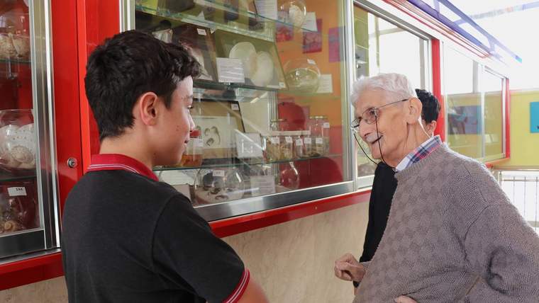 Bir öğrenci, sergi vitrininde bulunan biyolojik örnekleri inceliyor, yanında bir yaşlı adam sohbet ediyor.