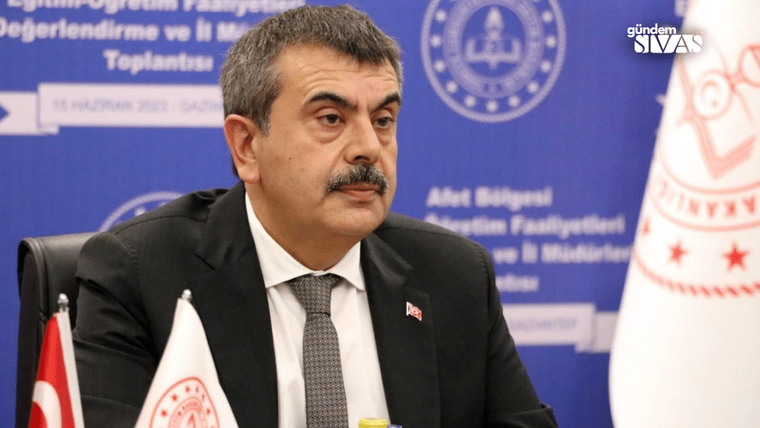 Masa başında oturan bıyıklı bir kişi, arkasındaki mavi toplantı fonu ve iki bayrakla görülüyor.