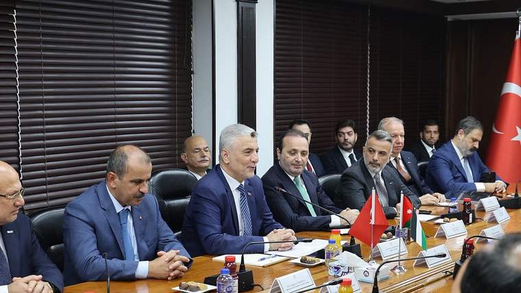 Toplantı masasında oturan kişiler, Türk ve Ürdün bayraklarının bulunduğu salonda konuşmayı dinliyor.