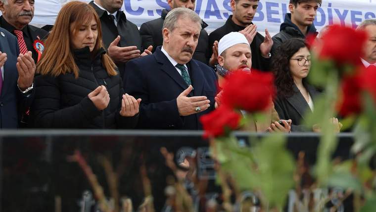 Bir grup insan, mezar başında dua ediyor; ön planda kırmızı güller var.