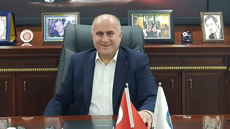 Bir kişi, ofisinde otururken gülümsemekte, arkasında çerçevelenmiş fotoğraflar var.