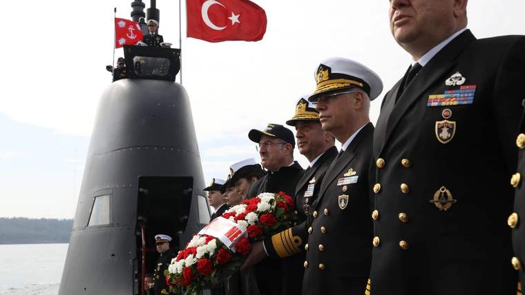 Denizaltı önünde Türk bayrağı ve çelenk taşıyan üniformalı kişiler, tören sırasında yan yana duruyor.