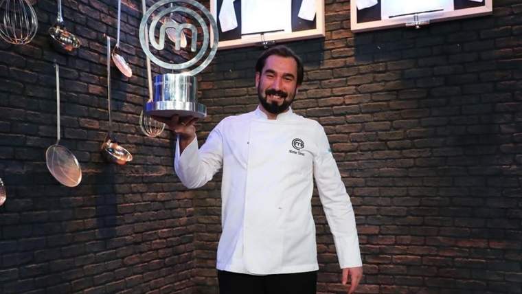 Beyaz aşçı ceketi giyen sakallı bir adam, elinde MasterChef logolu kupa tutuyor.