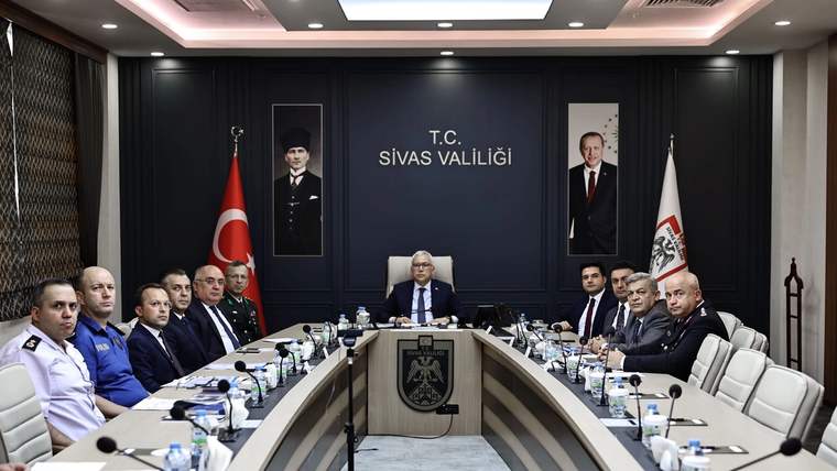 Toplantı salonunda uzun masanın çevresinde oturan kişiler, ortadaki yöneticiyi dinliyor; arkada Sivas Valiliği yazısı görülüyor.