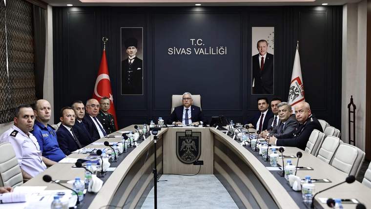 Toplantı salonunda uzun masanın çevresinde oturan kişiler, ortadaki yöneticiyi dinliyor; arkada Sivas Valiliği yazısı görülüyor.