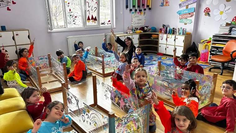 Renkli kıyafetler giymiş çocuklar, sınıfta el kaldırarak gülümsüyorlar.