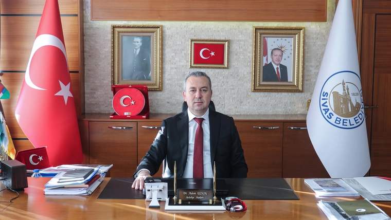 Bir kişi, Türk bayrağı ve üzerinde Sivas Belediyesi yazan bayrağın önünde makam masasında oturuyor.