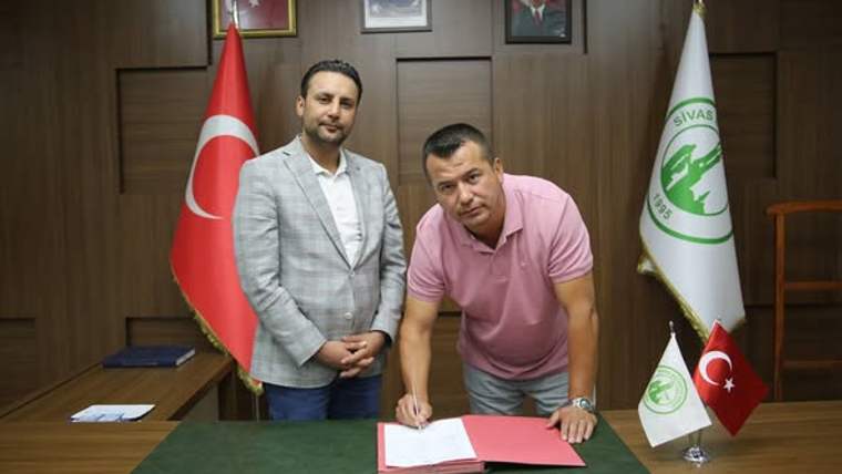 Masa başında iki erkekten biri belge imzalarken, arkalarında Türk bayrağı ve yeşil beyaz flamalar görülüyor.