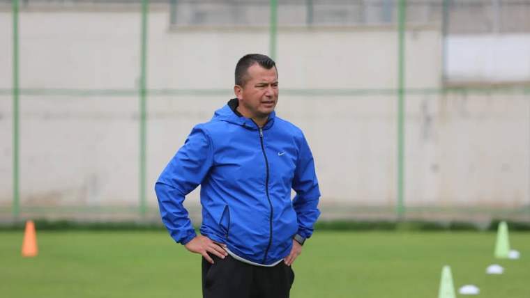 Mavi eşofman üstü ve siyah pantolon giyen bir kişi, futbol sahasında elleri belinde duruyor.