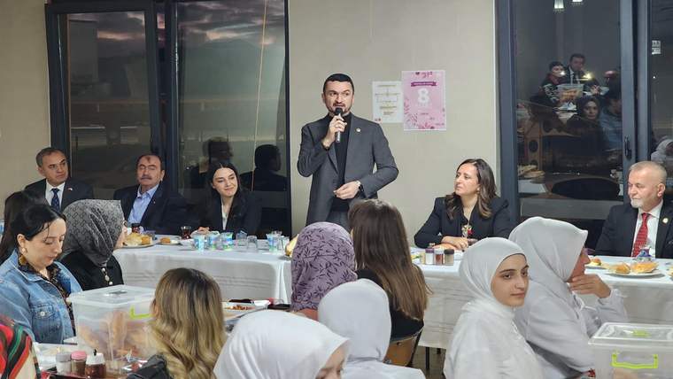 Mikrofonla konuşan bir kişi, yemek masalarının bulunduğu salonda oturan davetlilerin önünde duruyor.