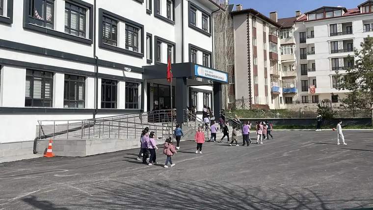 Bir binanın önündeki asfalt alanda çeşitli yaşlarda çocuklar duruyor; girişte Türk bayrağı görülüyor.