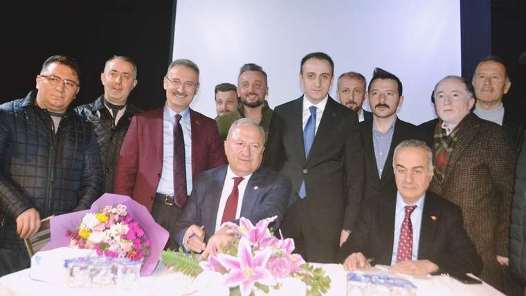 Masa önünde oturan iki erkek, arkalarında duran erkek grubuyla birlikte çiçeklerin arasında poz veriyor.