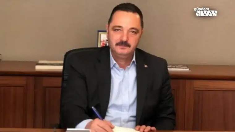Bir masa başında oturan, kalemle not alan bir adam, resmi bir kıyafet giymekte.