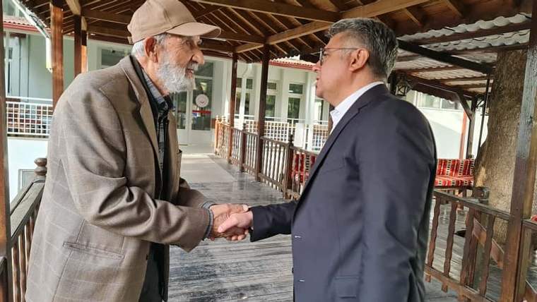 Ahşap sundurma altında yaşlı bir erkek ile takım elbiseli bir erkek tokalaşarak konuşuyor.