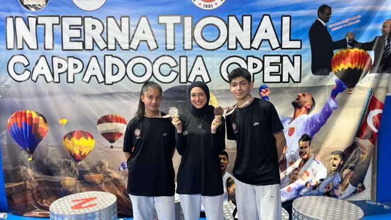 Üç genç, üzerinde “International Cappadocia Open” yazan pankartın önünde yan yana duruyor.