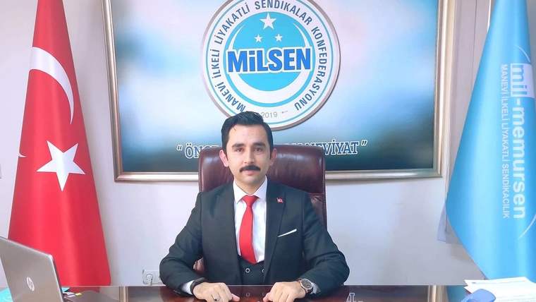 Masanın arkasında takım elbiseli bir kişi oturuyor; arkasında MİLSEN logosu, Türk bayrağı ve mavi bayrak bulunuyor.
