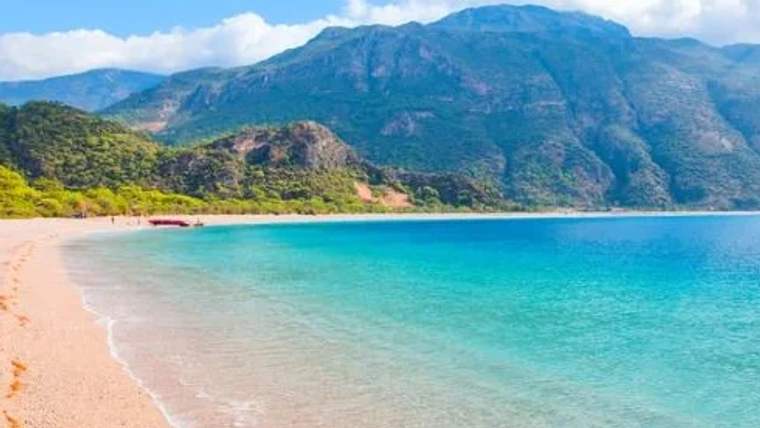 Sakin bir plaj, deniz ve arka planda dağlar görünmekte.