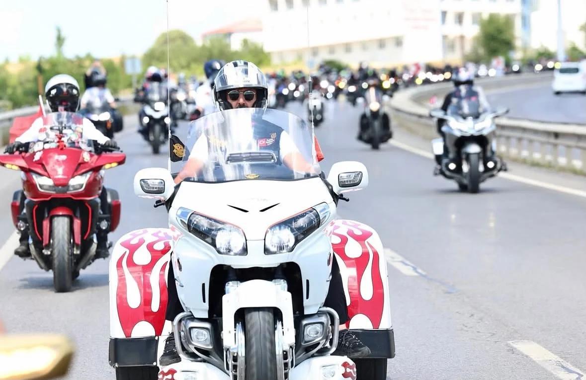 Kasklı sürücülerin bulunduğu motosiklet konvoyunda, önde beyaz ve kırmızı üç tekerlekli motosiklet ilerliyor.