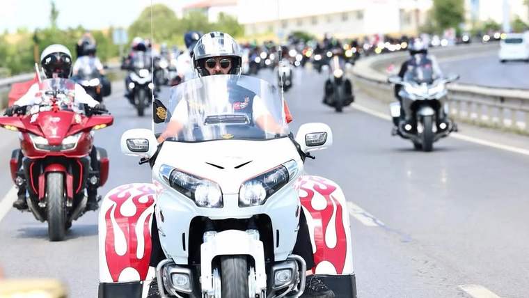 Kasklı sürücülerin bulunduğu motosiklet konvoyunda, önde beyaz ve kırmızı üç tekerlekli motosiklet ilerliyor.