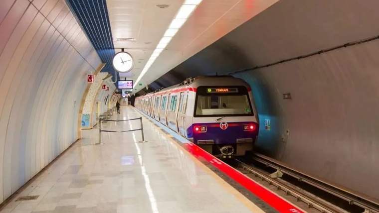 Yer altındaki istasyon platformunda kırmızı-beyaz bir metro treni raylarda duruyor; duvarda saat görülüyor.