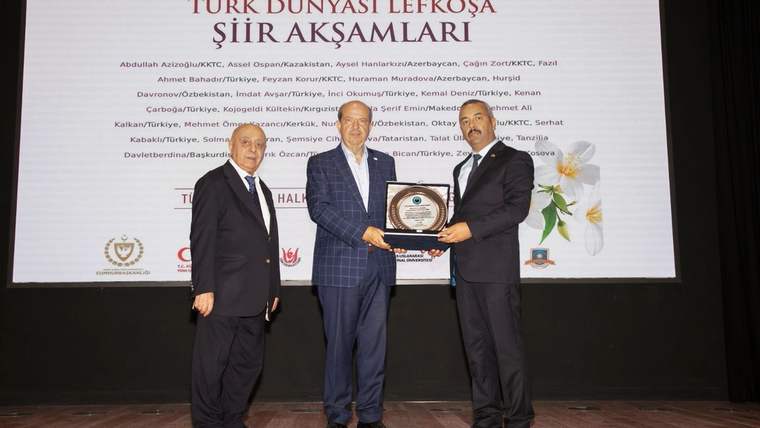 Sahnede üç kişi, ortadaki kişinin elinde plaketle duruyor; arkadaki perdede etkinlik yazıları yer alıyor.