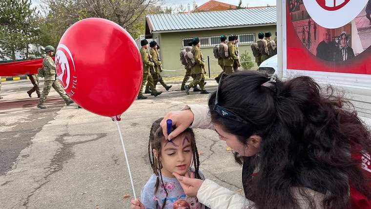 Bir kadın, elindeki kalemle küçük bir kızın alnına şekil çizerken kız kırmızı balon tutuyor; arkada askerler yürüyor.