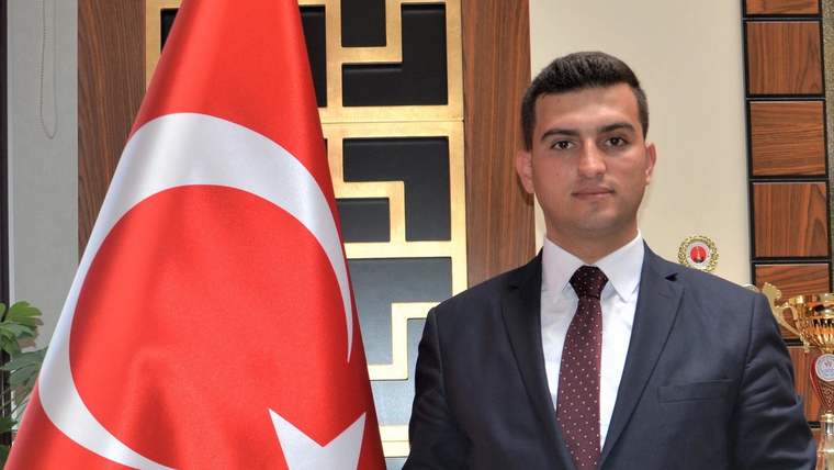 Bir kişi, arkasında Türk bayrağı ile resmi bir kıyafet içinde poz veriyor.