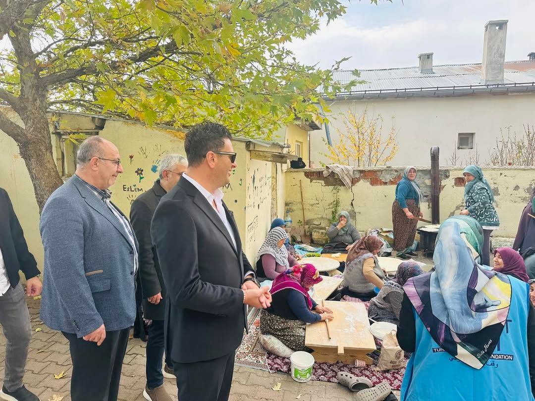 Ağaçlı bir avluda, masanın çevresinde oturan kadınlar ve yanlarında duran üç erkek görülüyor.