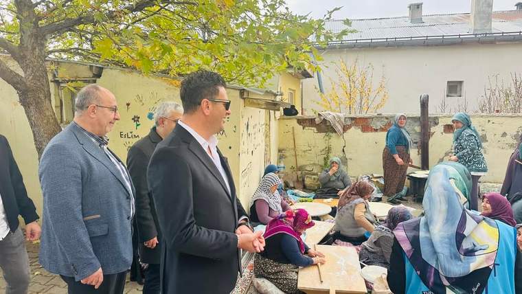 Ağaçlı bir avluda, masanın çevresinde oturan kadınlar ve yanlarında duran üç erkek görülüyor.