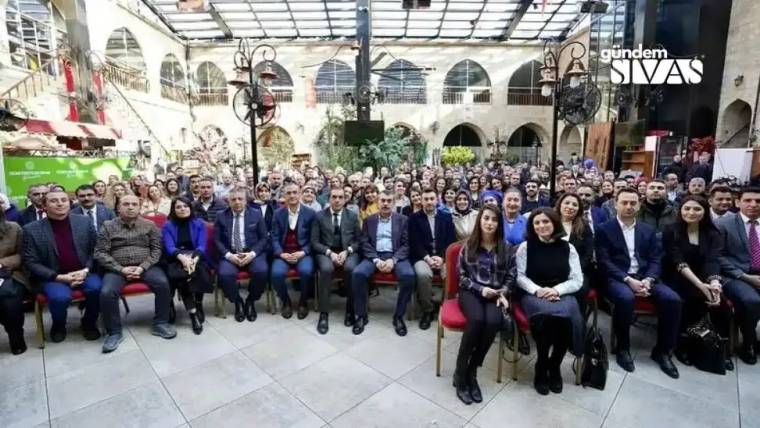 Bir etkinlikte oturan kalabalık bir grup insan, bazıları gülümseyerek kameraya bakıyor.