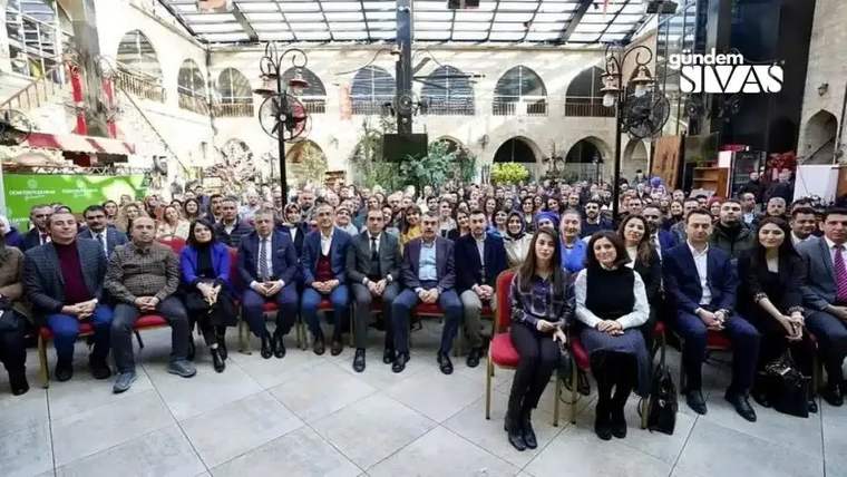 Bir etkinlikte oturan kalabalık bir grup insan, bazıları gülümseyerek kameraya bakıyor.