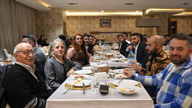 Restoran salonunda uzun bir masada oturan kalabalık grup, yemek tabakları ve içeceklerle görülüyor.