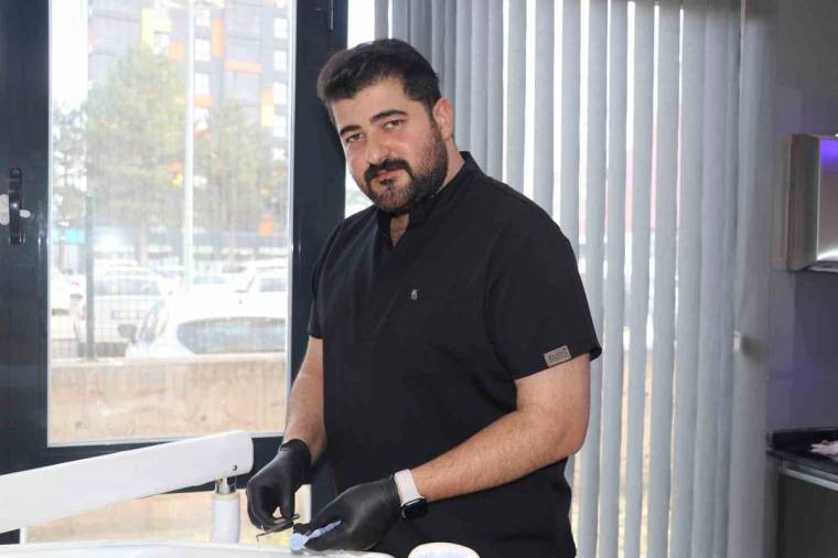 Siyah giyimli ve eldivenli bir kişi, diş tedavisi koltuğunun yanında kameraya bakıyor.
