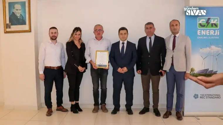 Bir grup insan, bir ödül belgesiyle birlikte poz veriyor. Arka planda bir afiş ve portre var.