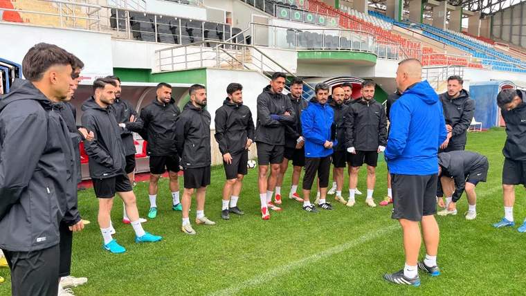 Futbolcular, stadyum tribünlerinin önündeki çim sahada mavi eşofmanlı bir kişinin çevresinde duruyor.