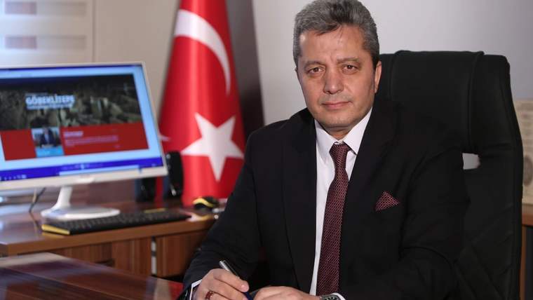 Takım elbiseli bir kişi masada oturuyor; arkasında Türk bayrağı, yanında bilgisayar ekranı bulunuyor.