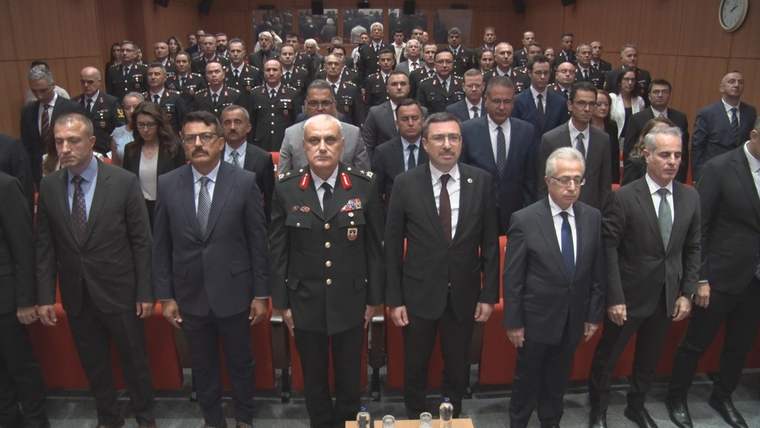Bir salonda üniformalı ve takım elbiseli çok sayıda kişi, sıralar halinde ayakta duruyor.
