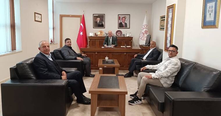Makam odasında Türk bayrağı ve kurum flamaları önünde, masanın çevresinde oturan beş kişi görülüyor.