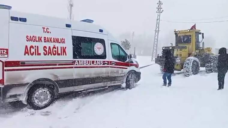 Karla kaplı bir yolda, bir iş makinesi ambulansı çekiyor. Kar yağışı yoğun.
