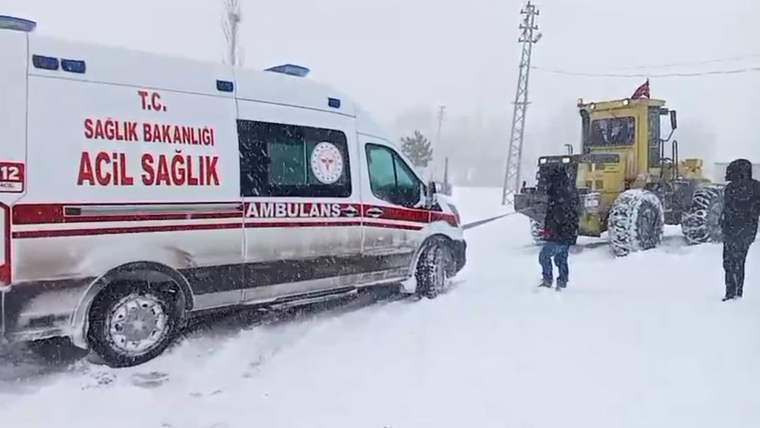 Karla kaplı bir yolda, bir iş makinesi ambulansı çekiyor. Kar yağışı yoğun.