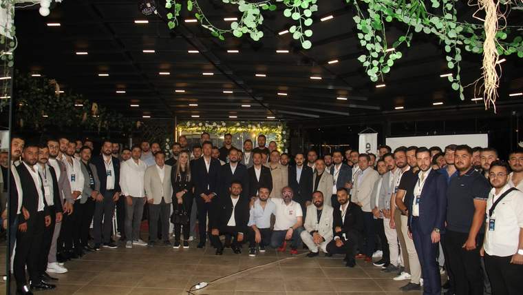 Kapalı bir salonda, asılı yeşil bitkilerin altında çok sayıda kişi birlikte poz veriyor.