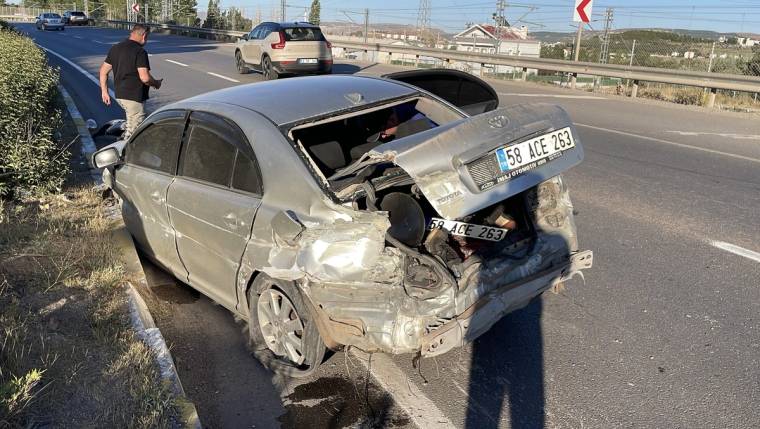 Yolda bariyerlere çarpmış bir araç, arka kısmı hasar görmüş. Yakınında birkaç kişi duruyor.