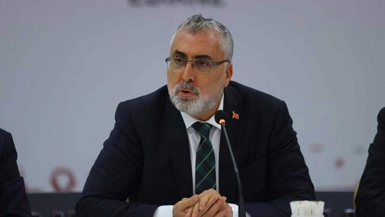 Koyu takım elbise giymiş, sakallı bir kişi, mikrofonun önünde konuşuyor.